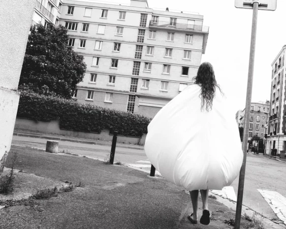 Person wearing a voluminous white garment walks down a city street, framed by urban buildings and trees in black and white.