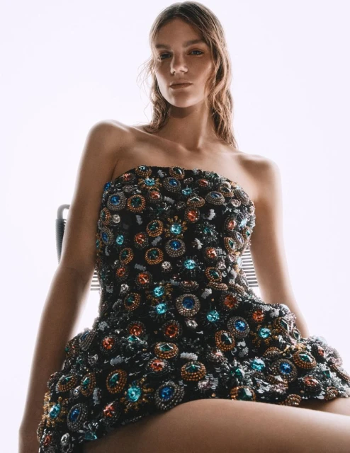 A woman with wavy light brown hair poses in a jeweled strapless minidress in a studio. The dress features colorful gems.