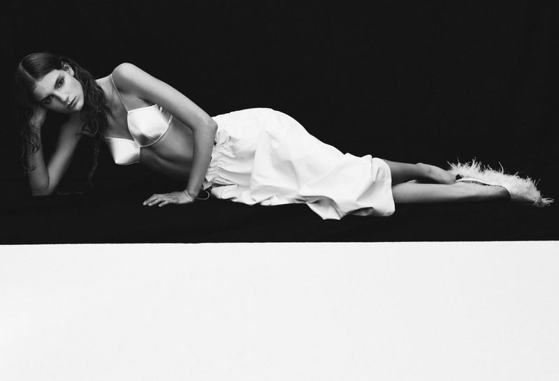 A young adult woman with long brown hair lies in repose wearing a white bra and skirt, photographed in black and white against a split background.