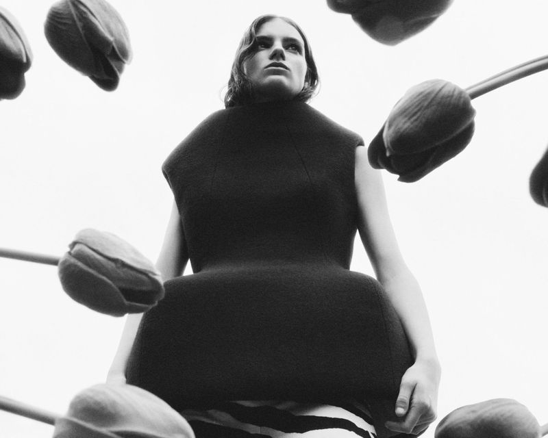 Model Sam Laban wearing a sculptural black structured top surrounded by tulip stems against a bright overcast sky.
