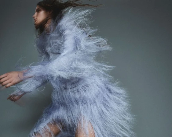 A fashion shot of a female model in motion, wearing a feathered dress, captured in a studio with a grey backdrop and dynamic blur.
