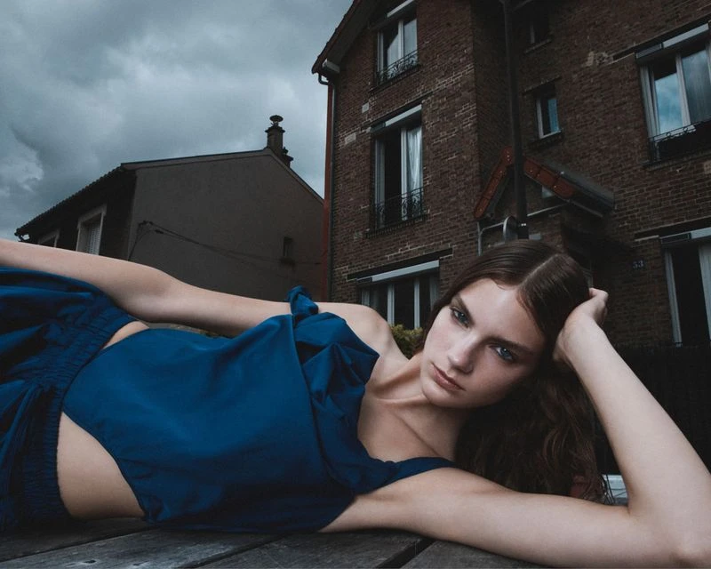 A woman poses in a blue dress outside, reclining on a wooden surface with brick buildings and an overcast sky in the background.