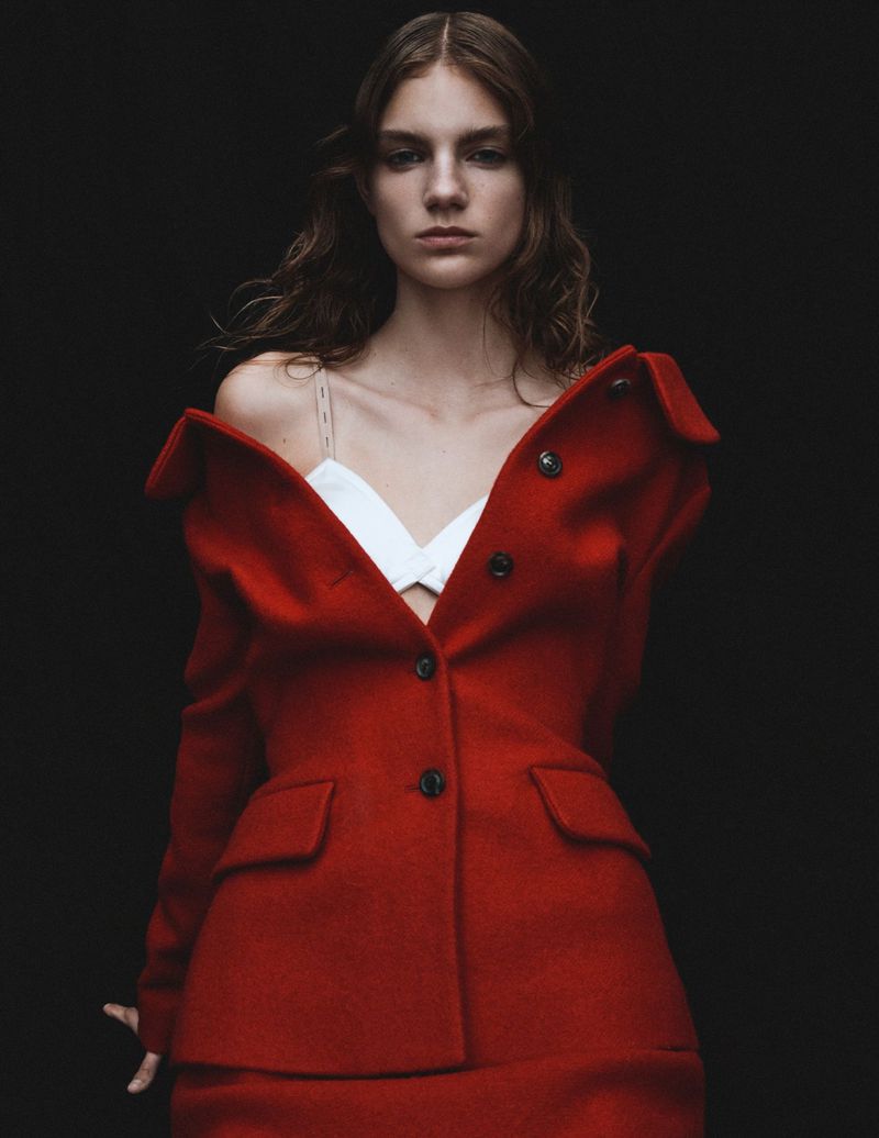 A woman with wavy dark hair stands against a black backdrop wearing a red blazer and skirt with a white top, in a studio shot.