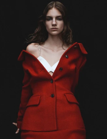 A woman with wavy dark hair stands against a black backdrop wearing a red blazer and skirt with a white top, in a studio shot.