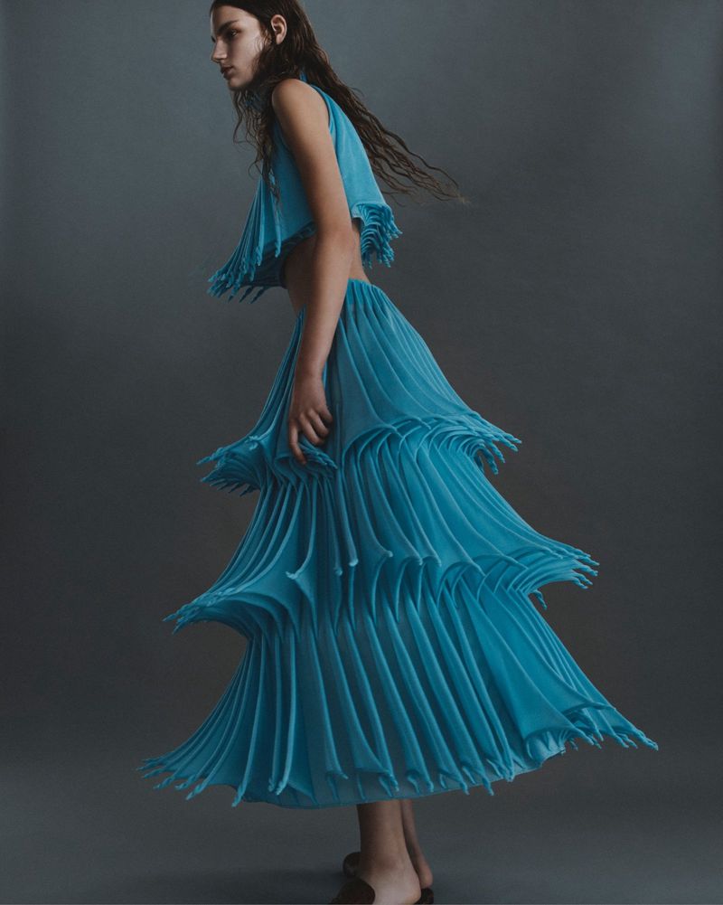 Fashion shot of a woman in a blue sleeveless top and a tiered, pleated skirt, posing in a studio with a neutral gray background.