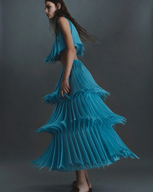 Fashion shot of a woman in a blue sleeveless top and a tiered, pleated skirt, posing in a studio with a neutral gray background.