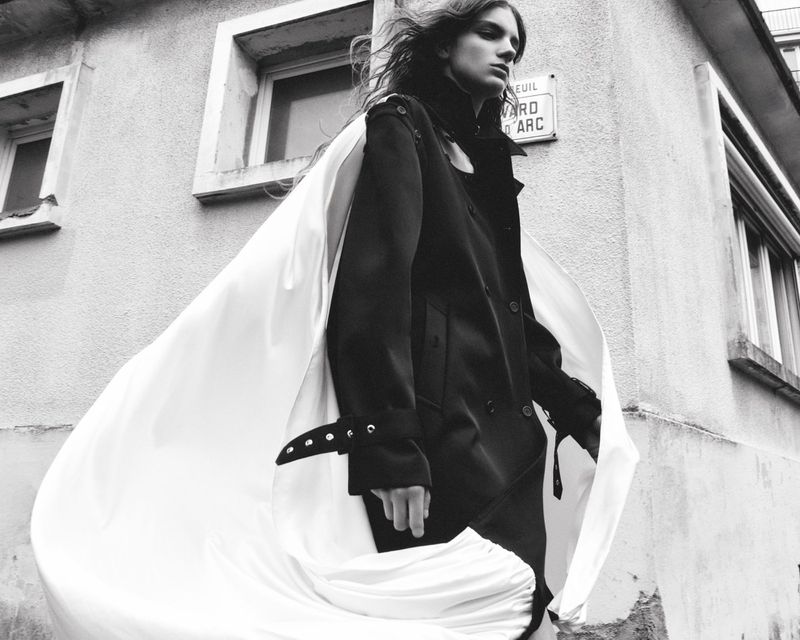 A stylish young adult in a dark coat and flowing white cape stands outside a light-colored building with windows, photographed in black and white.