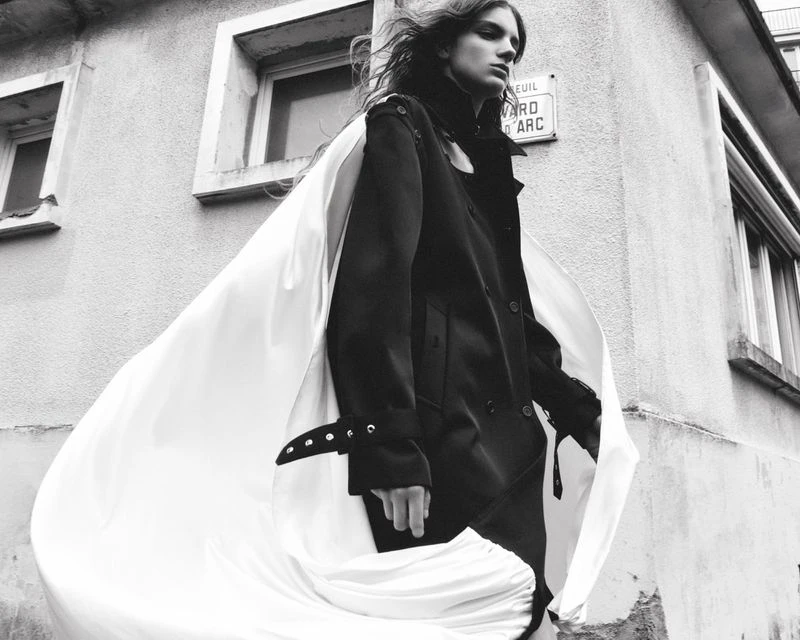 A stylish young adult in a dark coat and flowing white cape stands outside a light-colored building with windows, photographed in black and white.