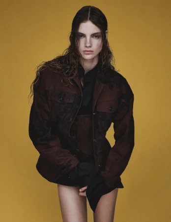Studio portrait of a young, thin brunette woman with wet hair wearing a dark jacket against a yellow background, looking at the camera.