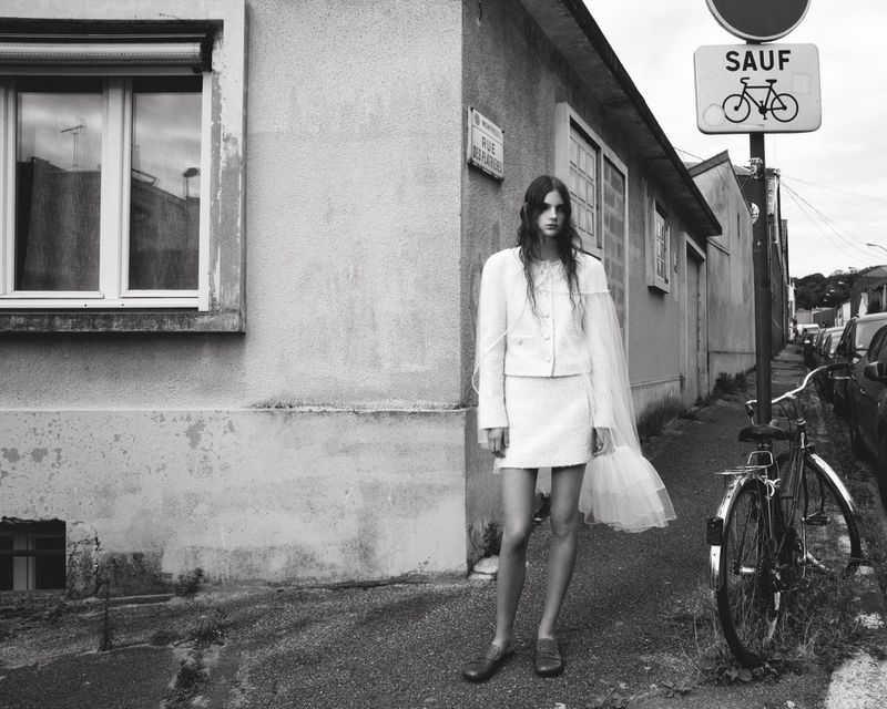 A woman in a white skirt suit stands outside on a sidewalk, near a bike and building, with natural light. The image is monochrome.