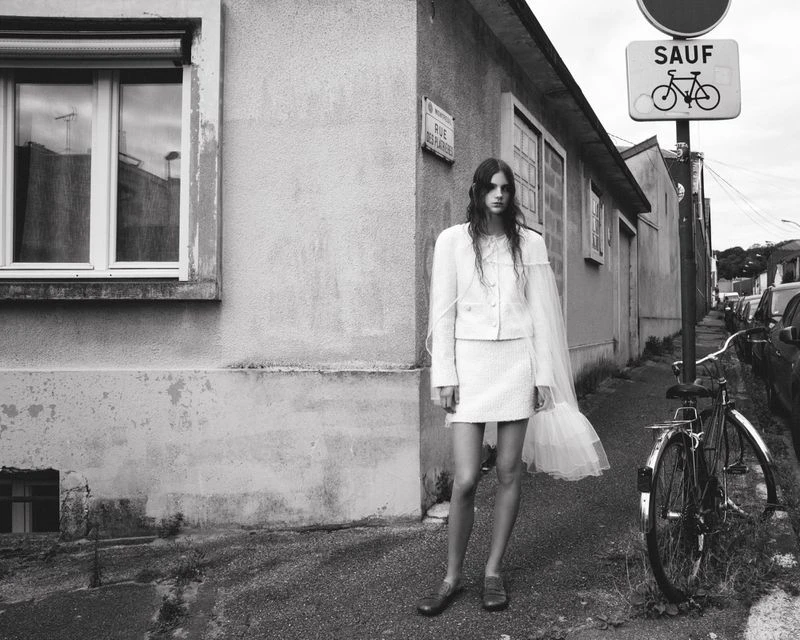 A woman in a white skirt suit stands outside on a sidewalk, near a bike and building, with natural light. The image is monochrome.
