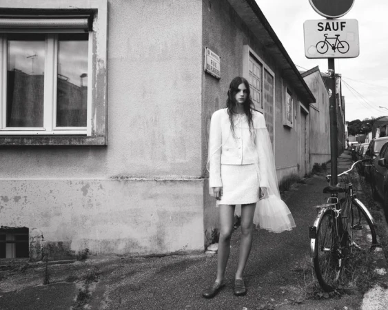 A woman in a white skirt suit stands outside on a sidewalk, near a bike and building, with natural light. The image is monochrome.