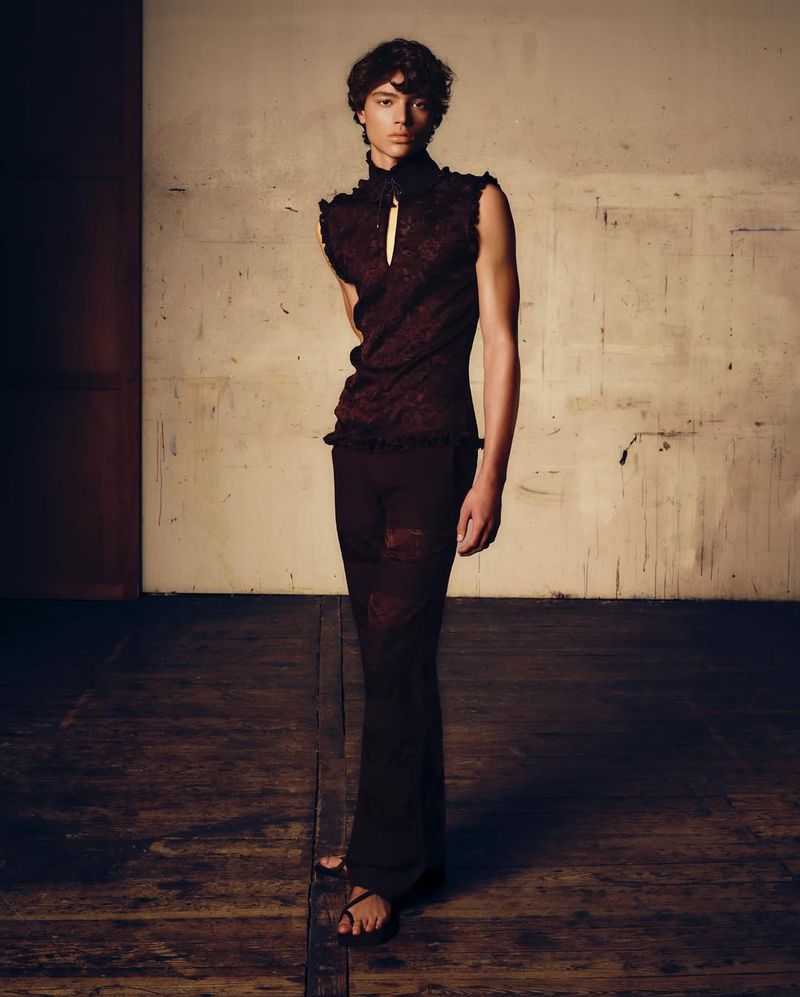 Full-length shot of an adult male model in a sleeveless burgundy top and flared trousers, posing in a photo studio against a grunge wall.