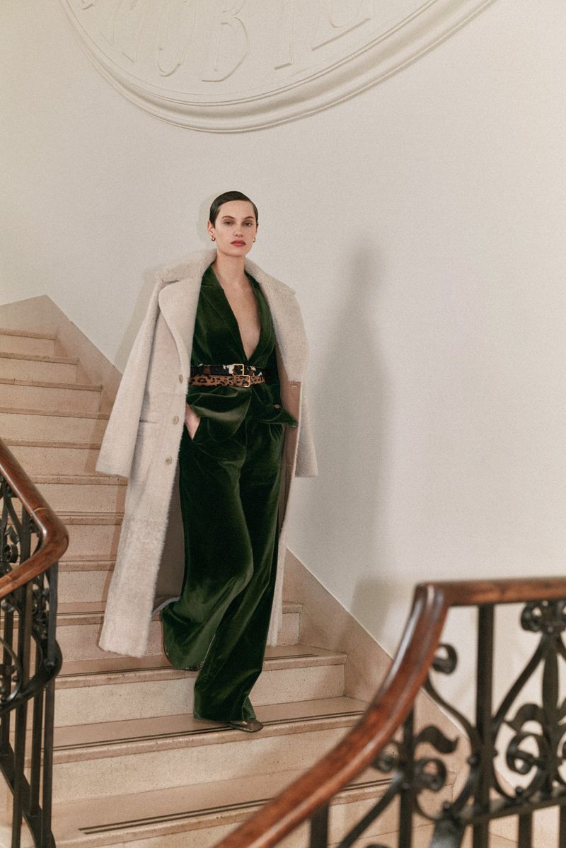 A woman wearing a green velvet suit and a long coat walks down a staircase in an indoor setting with white walls and a decorative railing.