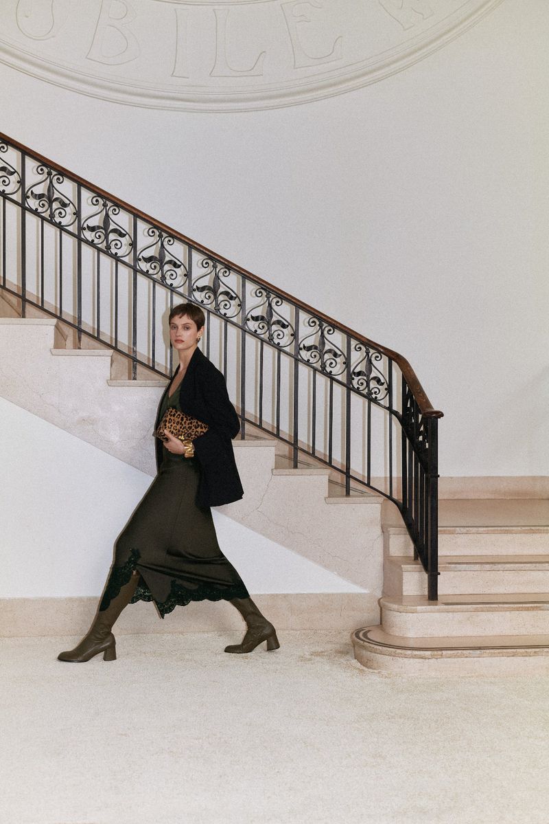 Woman in a dark green dress, black blazer, and olive green boots walks down marble stairs carrying a leopard print clutch.