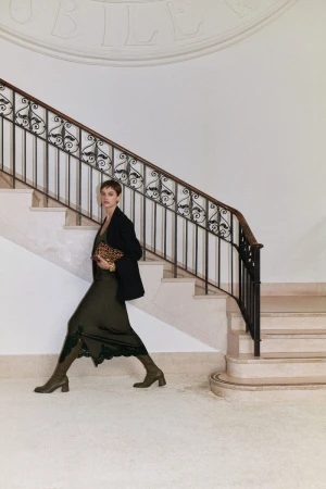 Woman in a dark green dress, black blazer, and olive green boots walks down marble stairs carrying a leopard print clutch.