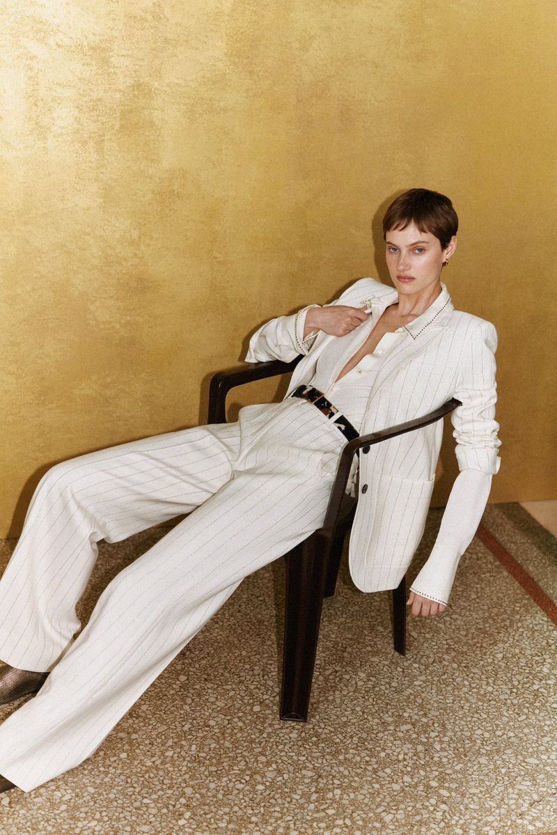 Fashion shot of a stylish woman in a pinstripe suit, sitting on a black chair against a gold backdrop with a neutral expression.