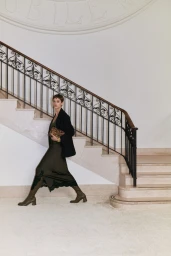 Model with short hair walking down grand indoor stairs wearing a dark coat and olive dress