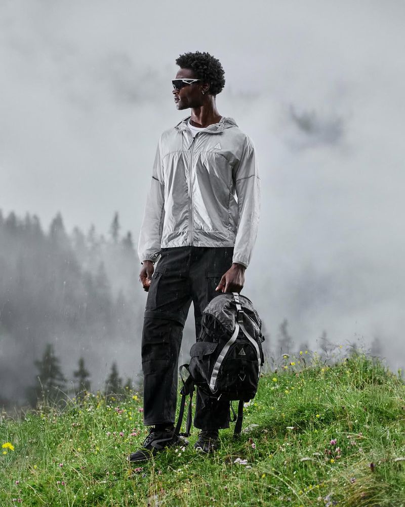 An African-American male model standing on a grassy hilltop in a light grey jacket and dark pants, holding a black backpack.