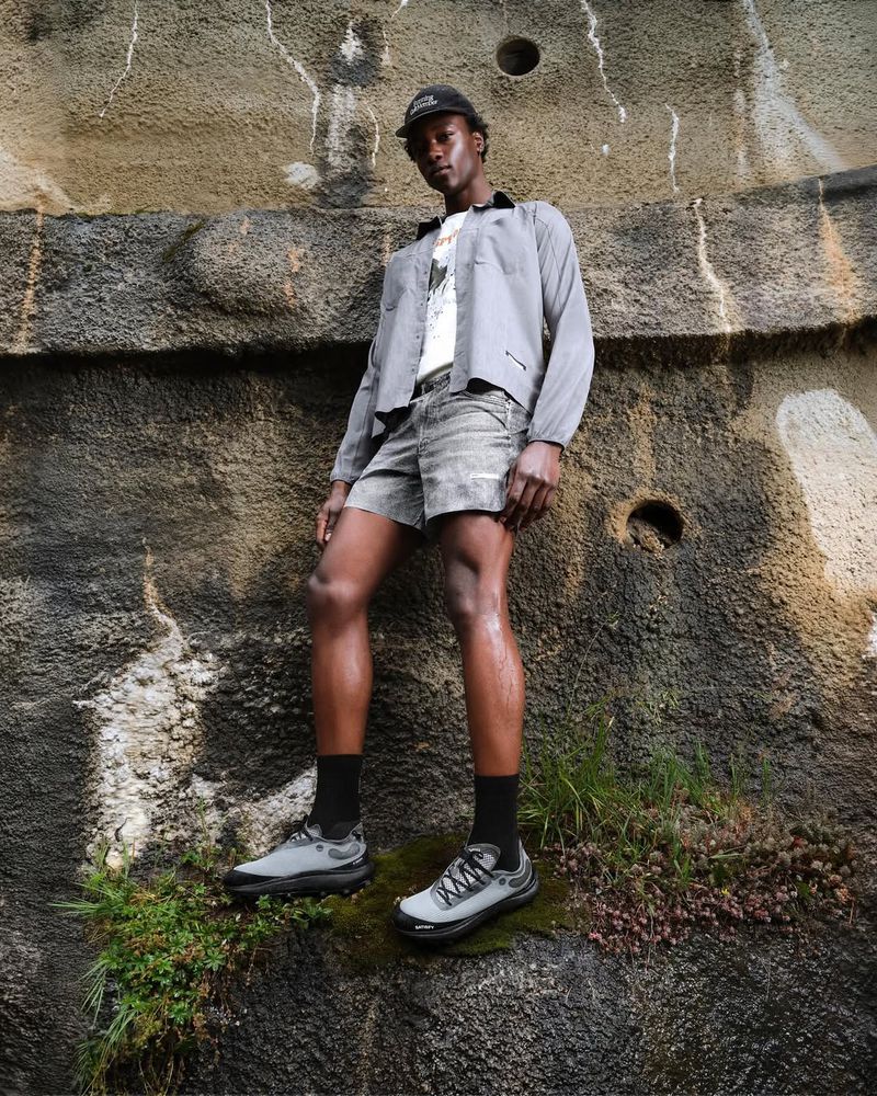 A man poses on a mossy rock formation wearing a gray jacket, patterned shorts, black socks, and gray running shoes in an outdoor setting.