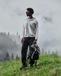 Black man wearing reflective windbreaker and dark cargo pants holding a backpack standing on a grassy hill