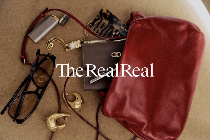 Still life of a burgundy leather purse, tortoiseshell hair comb, eyeglasses, and gold earrings on a tan surface with 'The RealReal' text.