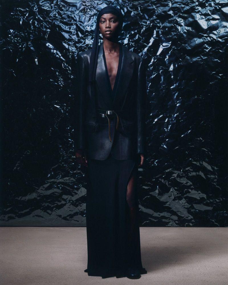 A woman stands in front of a textured black background, wearing a black wrap, blazer, and maxi skirt in a fashion editorial shot.