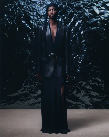 A woman stands in front of a textured black background, wearing a black wrap, blazer, and maxi skirt in a fashion editorial shot.
