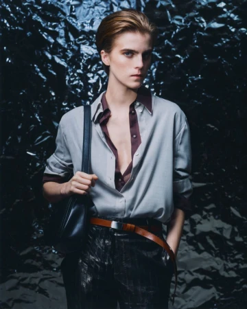 Fashion shot of an adult male in an unbuttoned shirt, brown belt, and pants, with a black shoulder bag against a dark backdrop.