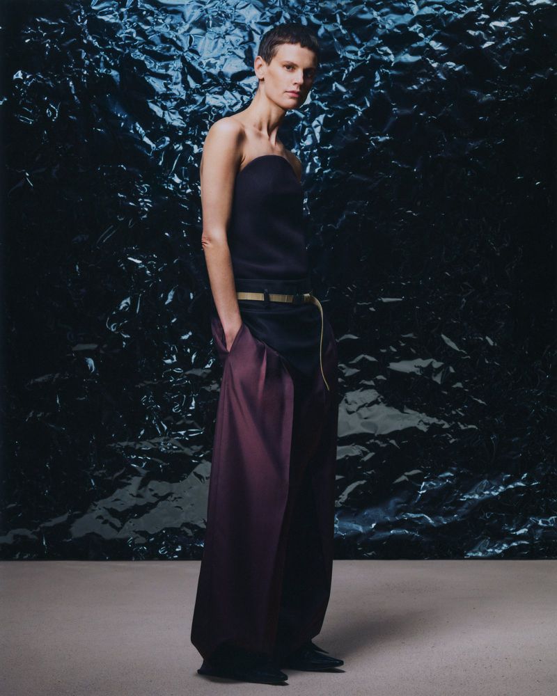 Model wears a strapless dark top with wide, dark and auburn pants against a dark, textured backdrop in a fashion studio setting.
