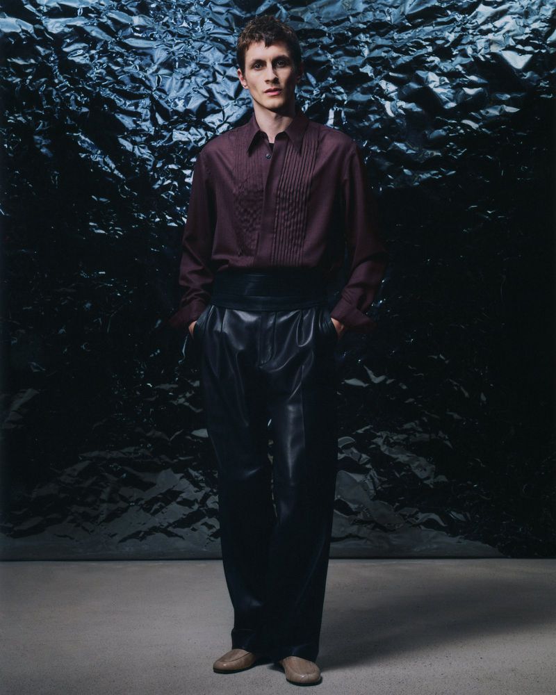 Man stands in a fashion pose wearing a dark red button-down shirt, black cummerbund, black leather pants, and light brown shoes.