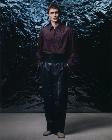 Man stands in a fashion pose wearing a dark red button-down shirt, black cummerbund, black leather pants, and light brown shoes.