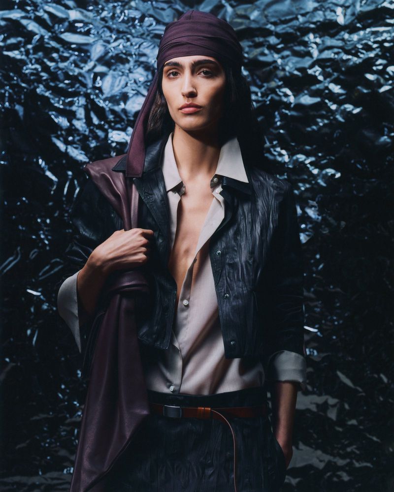 Fashionable adult female posing with a leather jacket slung over her shoulder, wearing a headscarf, blouse, skirt, and belt against textured background.