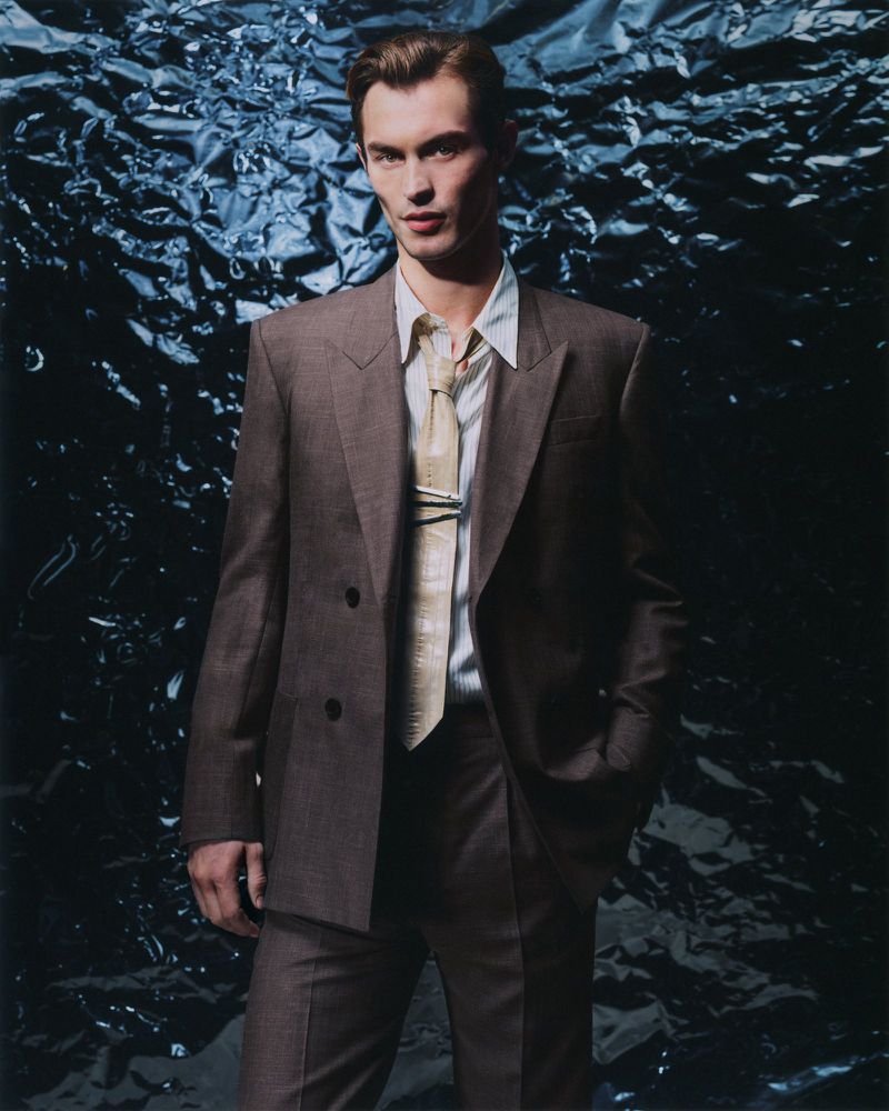 A man in a brown suit and light-colored tie stands in front of a crumpled blue foil background, captured in a studio-lit fashion shot.