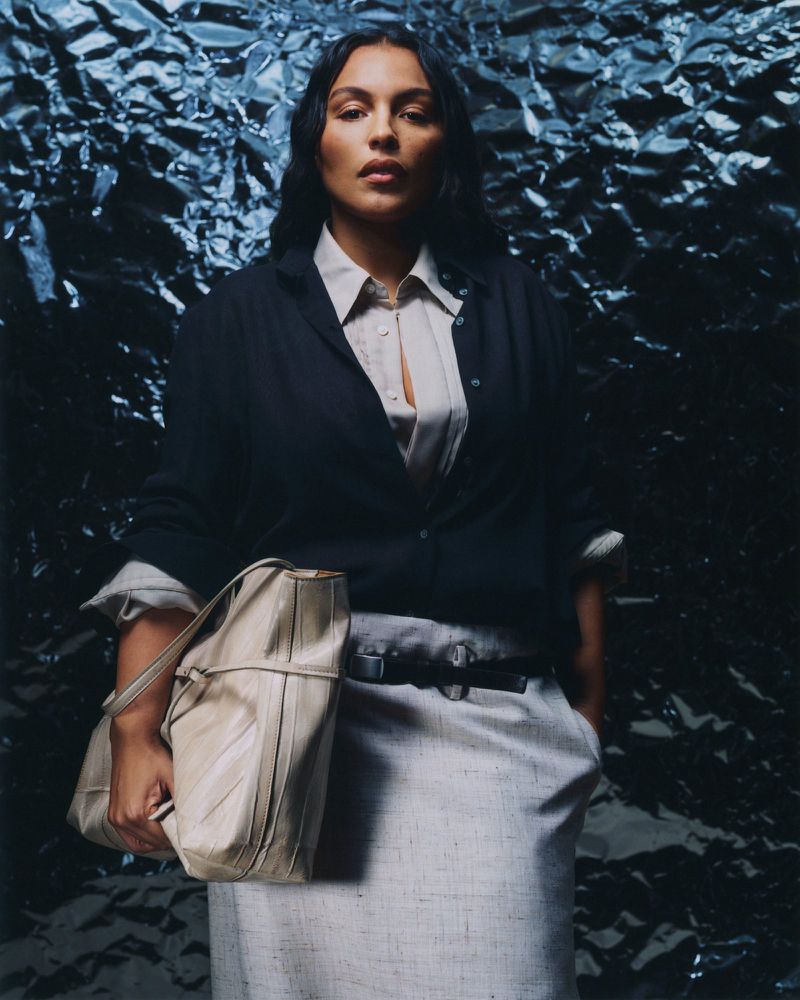A poised woman with long dark hair, wearing a black cardigan, light shirt and trousers, holds a beige bag against a textured dark backdrop.