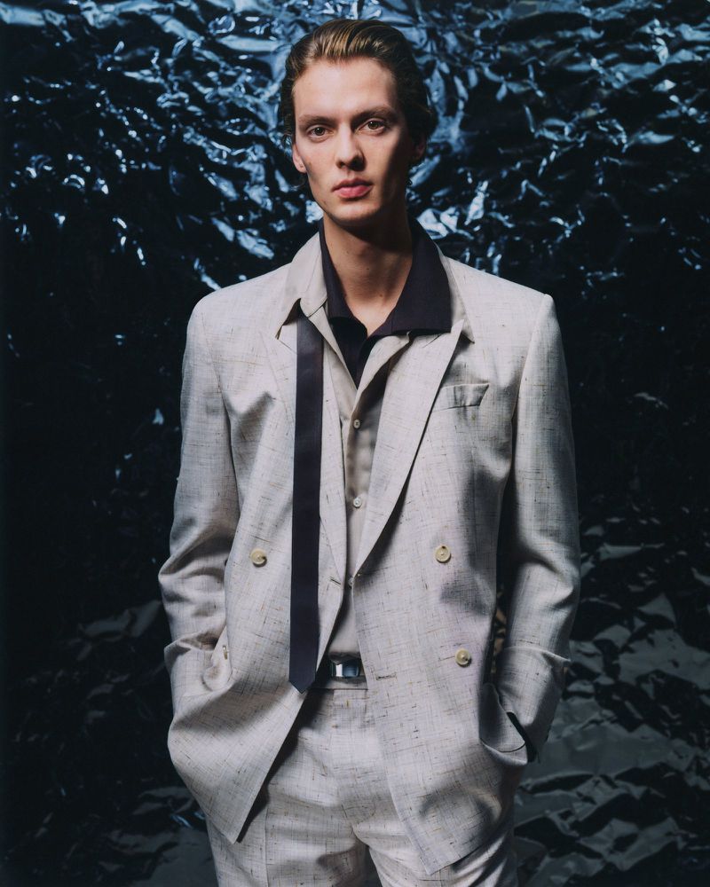 Man poses in a light-colored double-breasted suit with a navy collared shirt in front of a dark, textured backdrop in a studio shot.