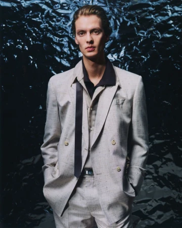 Man poses in a light-colored double-breasted suit with a navy collared shirt in front of a dark, textured backdrop in a studio shot.