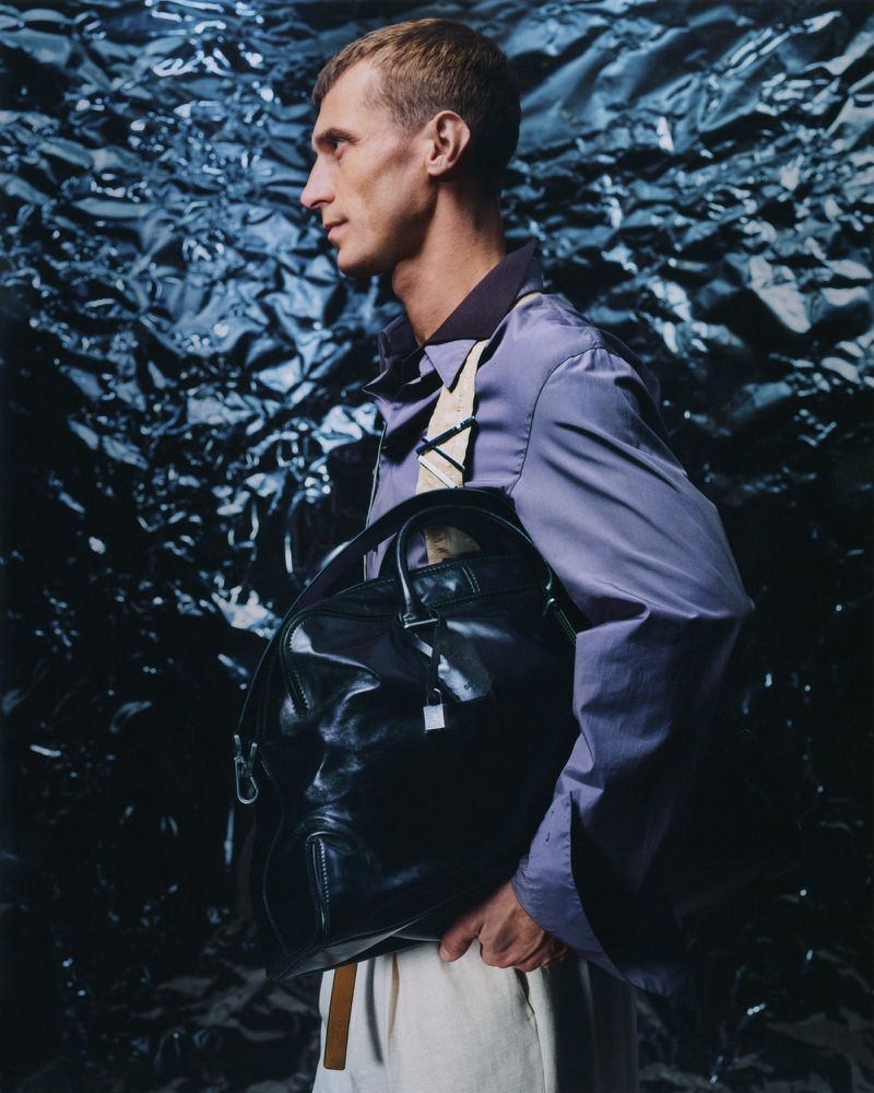 Profile shot of a man wearing a light purple jacket and white pants with a black bag, against a textured dark blue background.