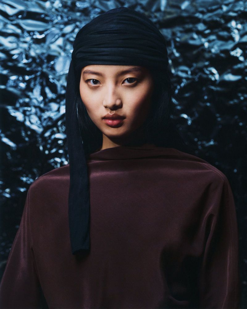 Portrait of an Asian woman wearing a black headscarf and a dark maroon blouse, set against a shimmering blue foil backdrop in a studio.