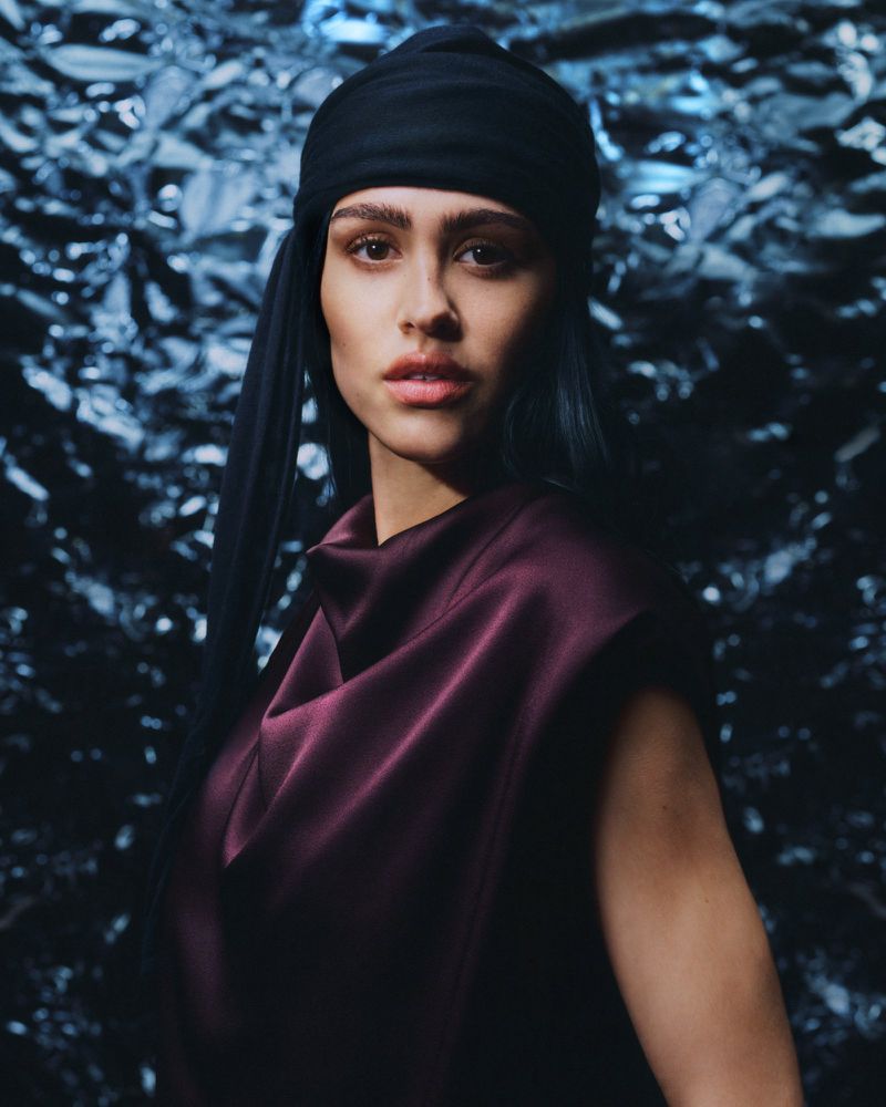Close-up portrait of a woman with a dark headscarf and burgundy top against a shiny, textured silver and blue background.