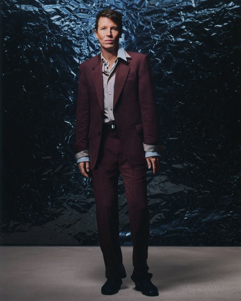 Full body shot of a man in a maroon suit against a dark blue crumpled foil background in a photo studio, showcasing fashion and style.