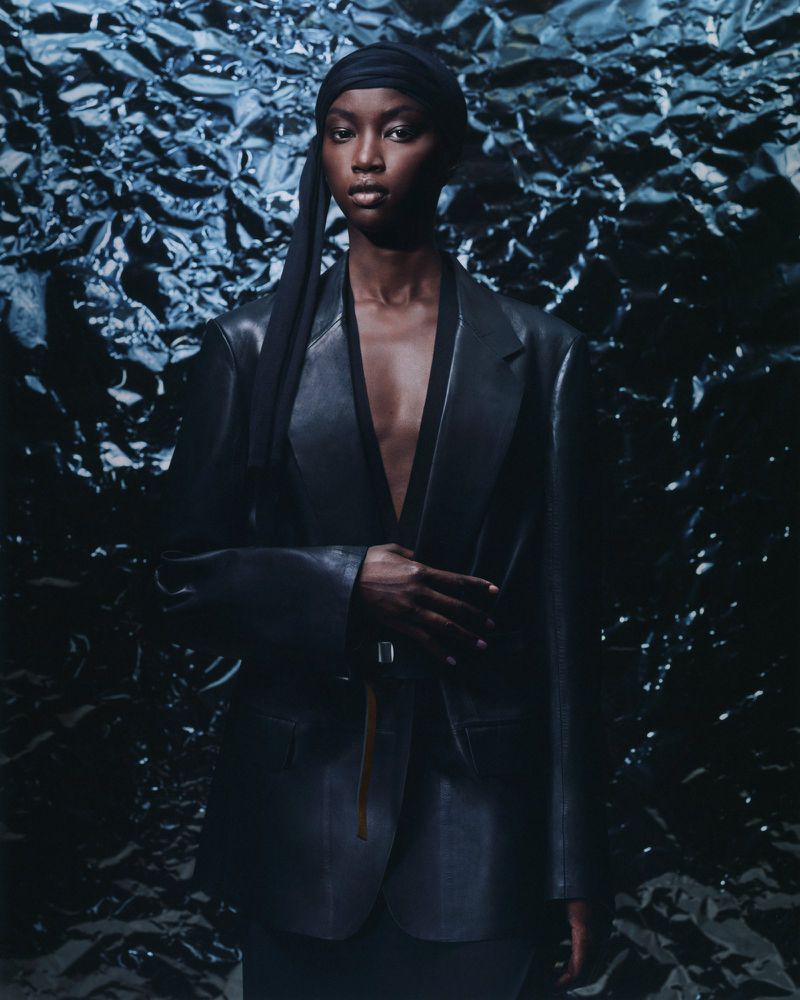 Portrait of a Black woman in an oversized leather blazer and headscarf, posing against a textured blue-gray background in a studio setting.