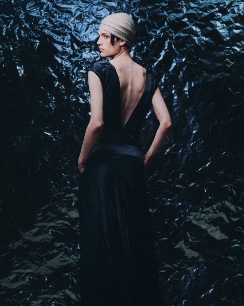 Elegant adult female in a dark dress and turban standing against a textured black background, photographed from behind.