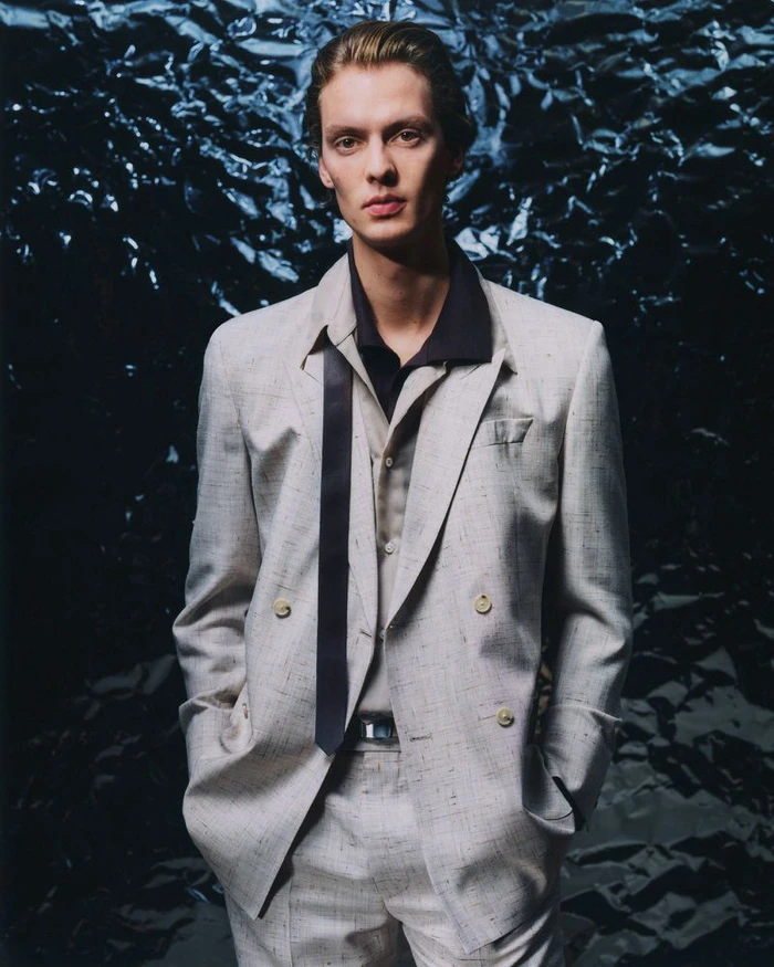 Portrait of a man wearing a light grey double-breasted suit and a dark untied necktie against a metallic textured foil background