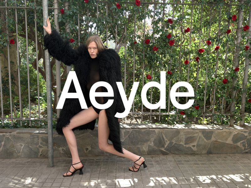 A fashion shot shows a woman in a shaggy coat and strappy heels posing on a sidewalk near a fence with red roses. The word 'Aeyde' is superimposed.