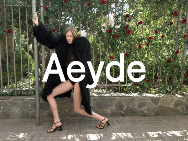 A fashion shot shows a woman in a shaggy coat and strappy heels posing on a sidewalk near a fence with red roses. The word 'Aeyde' is superimposed.