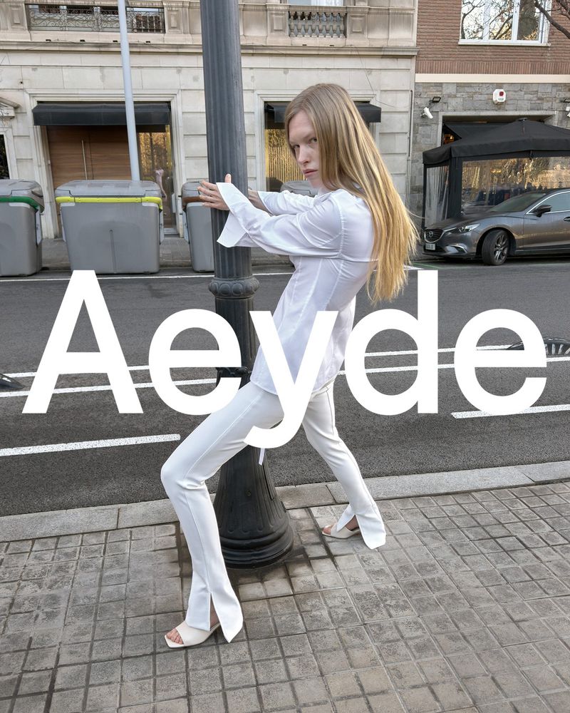 Blonde adult posing in a white pantsuit against a city street scene with building facades, streetlights, and a grey car.
