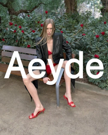 Fashion editorial shot of a slender adult female model in a black leather jacket and red shoes, posed on a park bench with white Aeyde text.