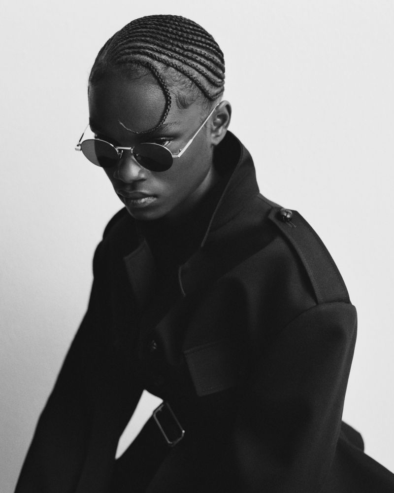 Fashion portrait of a young African woman wearing sunglasses and a black wool coat against a light gray backdrop in a studio.