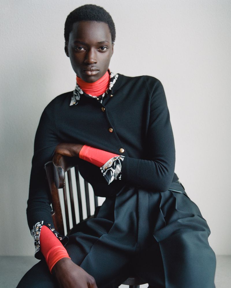 Portrait of a Black woman in a black cardigan and wide-leg trousers sitting on a chair against a plain wall.